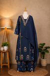 A navy blue three-piece salwar kameez suit set with colorful embroidered patterns and gold zari detailing, displayed on a mannequin in a styled room setting with a floor lamp, plants, and a wooden stool.