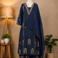 A navy blue three-piece salwar kameez suit set with colorful embroidered patterns and gold zari detailing, displayed on a mannequin in a styled room setting with a floor lamp, plants, and a wooden stool.